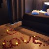 Chambre romantique avec le mot 'LOVE' formé en pétales de roses rouges sur le sol, entouré de bougies, avec un lit élégant en arrière-plan