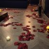 Chemin de pétales de roses rouges et bougies allumées sur un sol en bois clair menant à un lit sombre dans une chambre romantique.